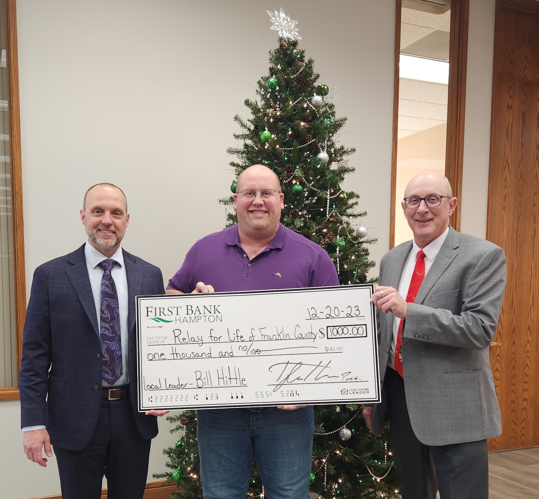 three people pose with a check from bank donation in front of Christmas tree.